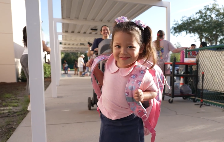 a little girl wearing a pink shirt and backpack and pigtails.