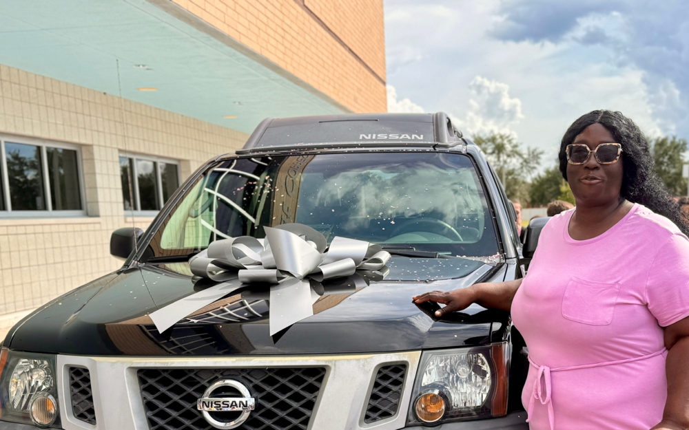 A Nissan with a silver bow on top was gifted to an employee wearing a pick dress standing next to it.
