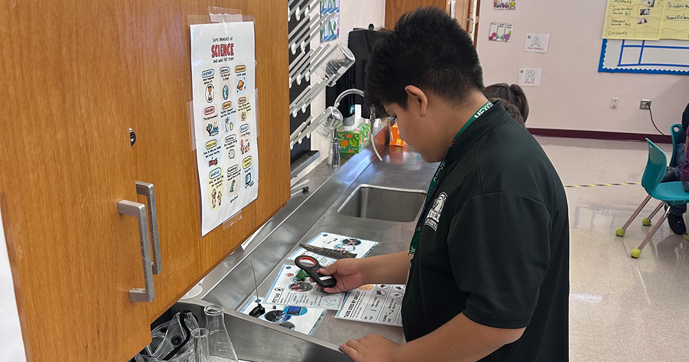 A student holds a magnifying glass up to learning materials in a classroom. 