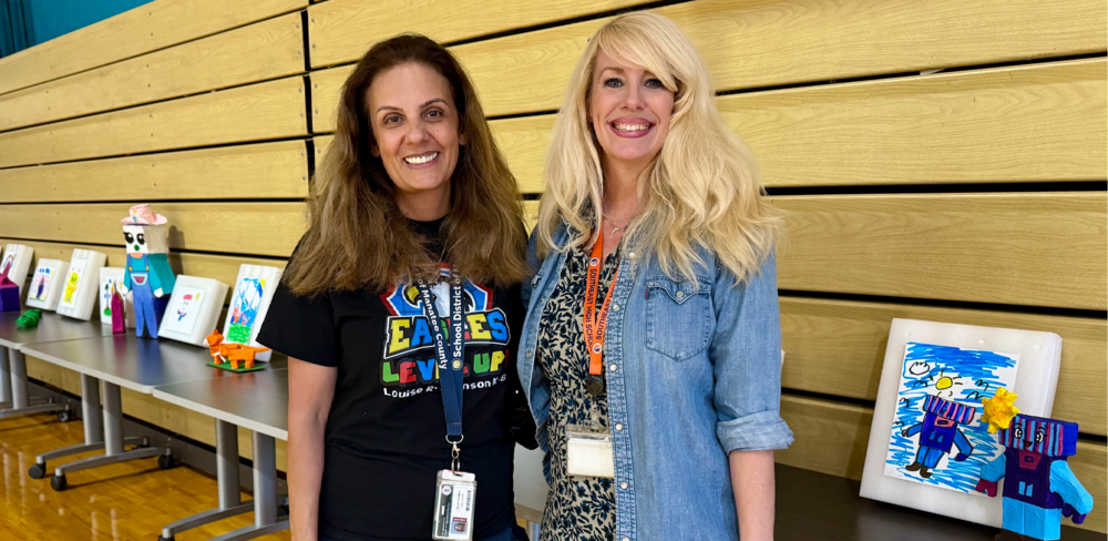 Two women smiling while standing in front of their student's artwork.