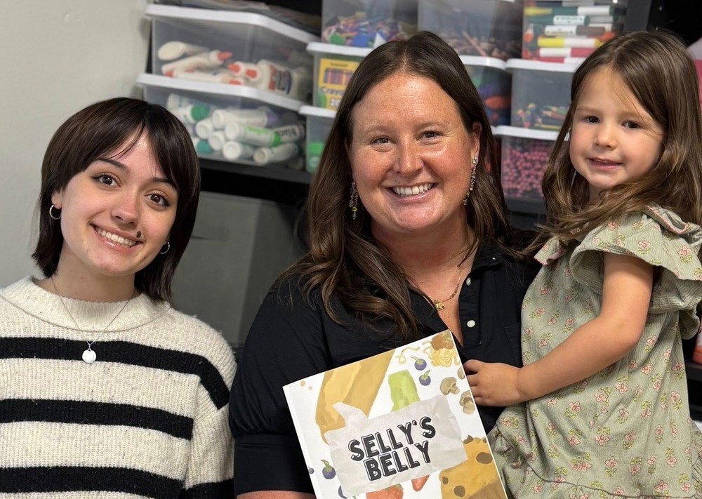 A teacher holding her daughter and first published book next to the former student who illustrated it.
