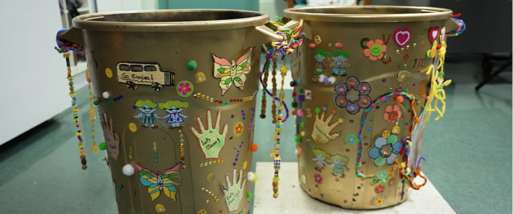 Two gold trash cans decorated with beads and school related drawings.