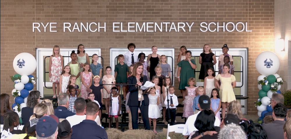 Students stand on chorus risers in front of a brick wall with a sign reading Rye Ranch Elementary School.