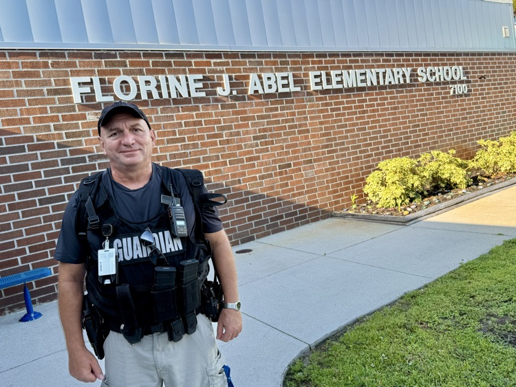 A school safety guardian wearing a vest stands in front of a school made from bricks