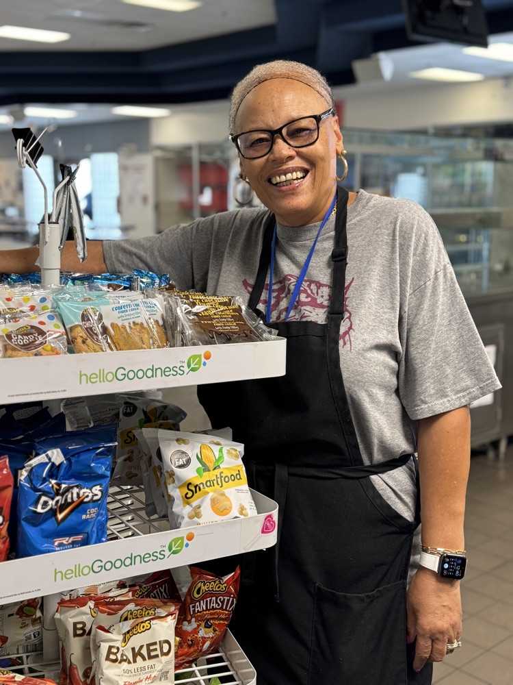 A smiling food service employee standing next to a rack filled with small bags of chips