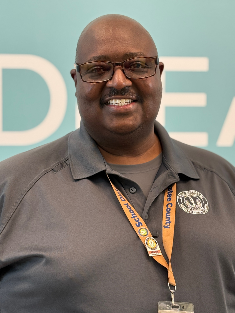 A school safety guardian wearing a bronze ID lanyard smiles at an elementary school.