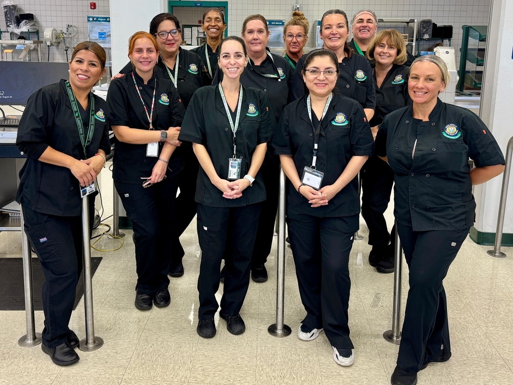A group of women and a man wearing black uniforms smiling in a high school cafeteria.