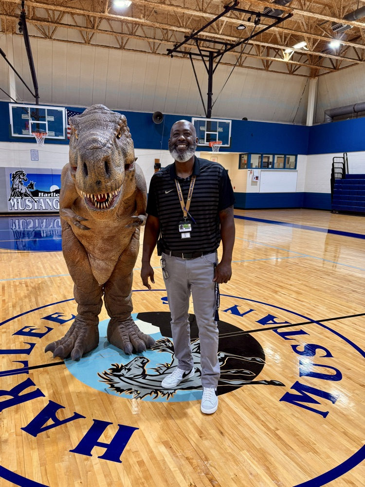 The principle of Harllee smiles while standing next to an animatronic dinosaur