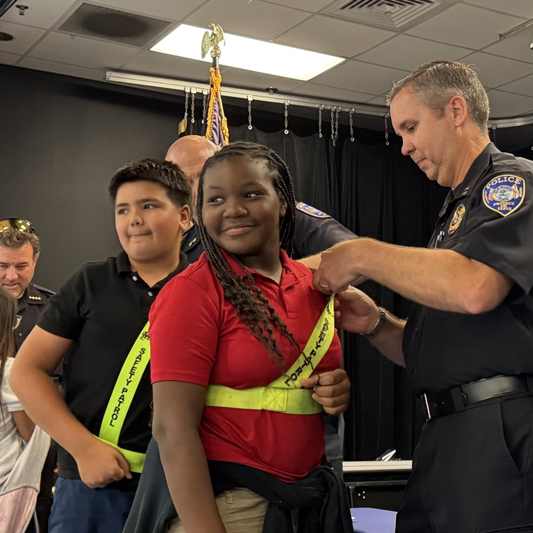 A student wearing a red shirt and safety patrol belt smiles as a police officer puts a pin on the belt