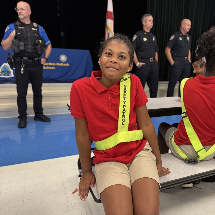A girl wearing a red shirt and bright yellow safety patrol belt smiles at a pinning ceremony
