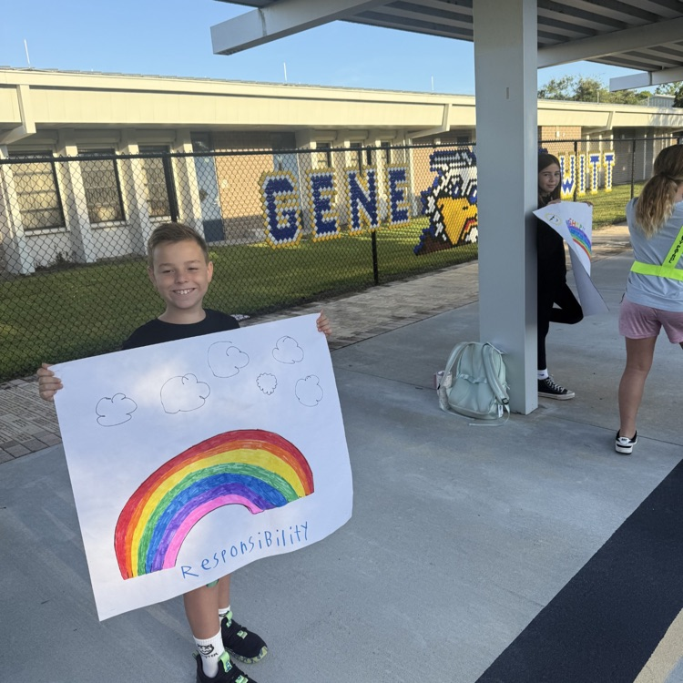 Students with signs stand on the sidewalk at school, waiting for peers to arrive and to say hello