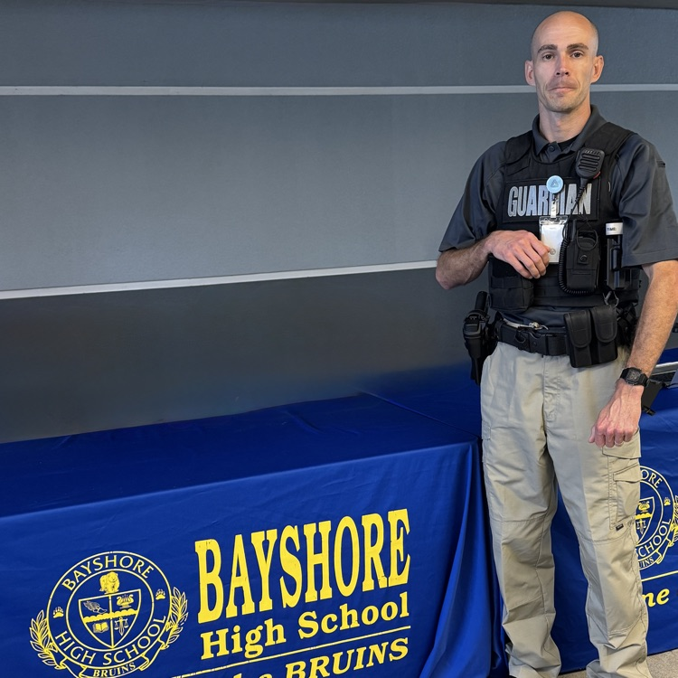 A school safety guardian standing next to a table covered in a blue tablecloth that says Bayshore high school home of the Bruins