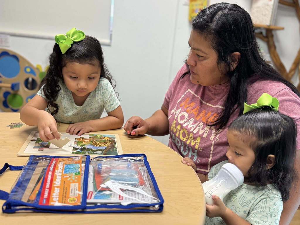 A mother and young girl look on as another girl puts together a puzzle.