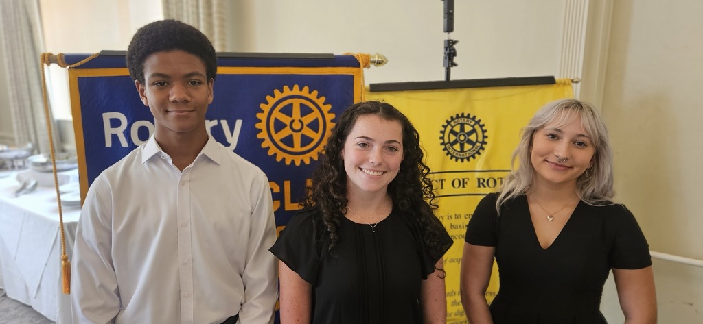 Three student smiling while standing in front of Rotary Club banners.
