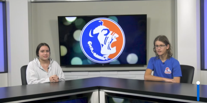Two student news anchors with a orange and blue high school logo for Southeast High in the background.