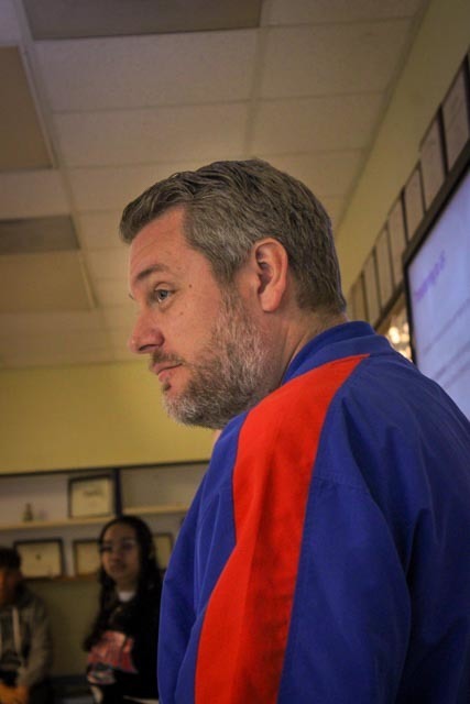 A man wearing an orange and blue jacket with a student looking on in the background.