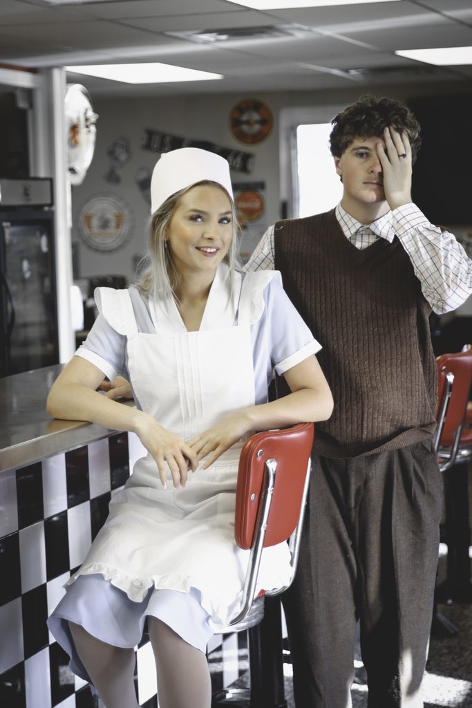 Two theatre actors in a diner. A young woman sitting and a young man holds a hand over his left eye.