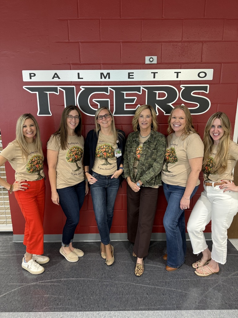 A group of women wearing the same shirts stand against a wall painted with the words, "Palmetto Tigers".