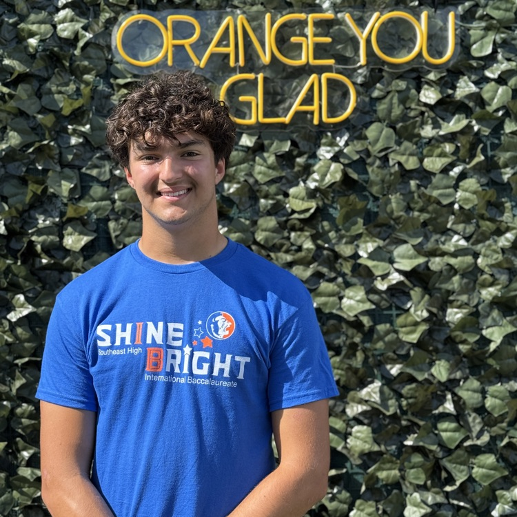 A young man wearing a blue school shirt stands in front of a back drop that says “orange you glad"