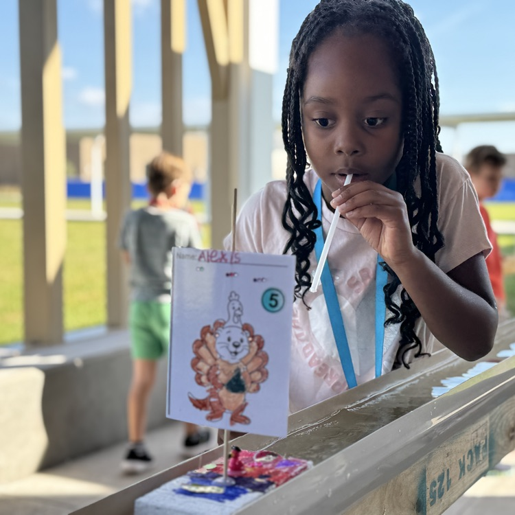 A young girl blows through a straw to move her handmade boat down a rain gutter