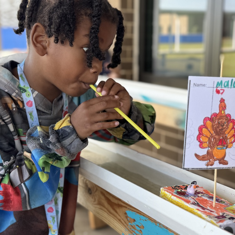 A young boy blows through a straw to move his handmade boat down a rain gutter