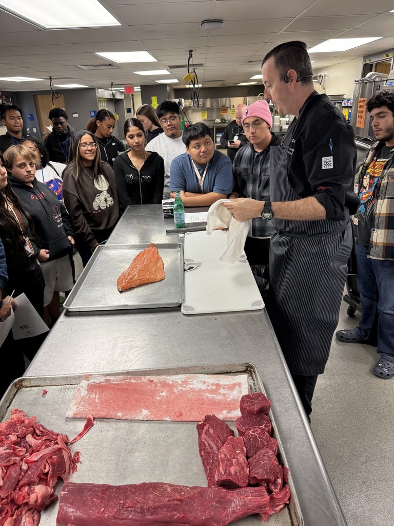 A chef doing a cooking demonstration in a culinary classroom at a high school with raw meat on the table