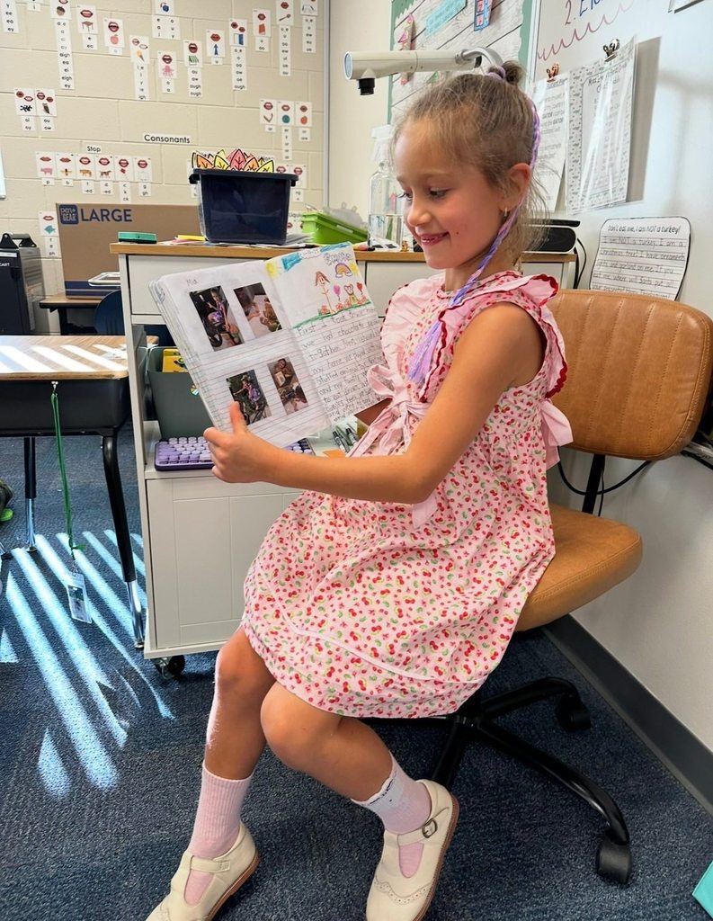 A young girl in a pink dress reads to classmates.