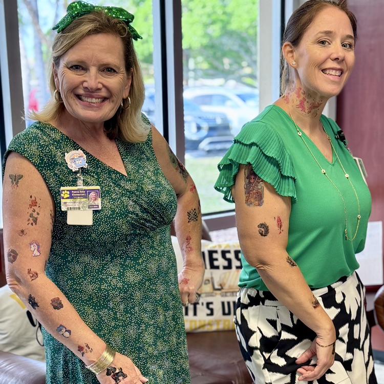 Two school administrators wearing green and smiling while showing off their temporary tattoos