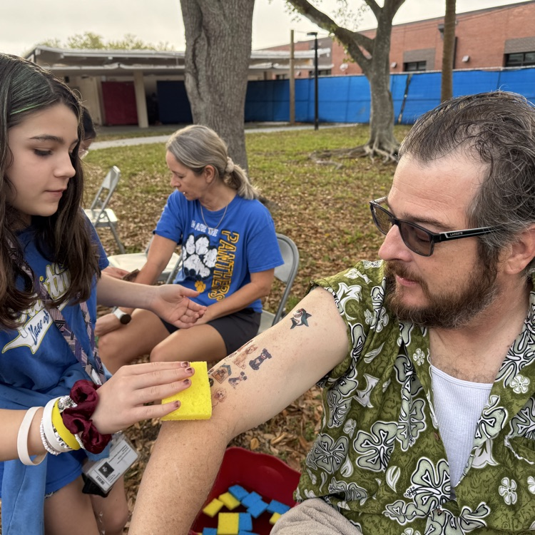 A male teacher holds out his arm while a student applies a temporary tattoo