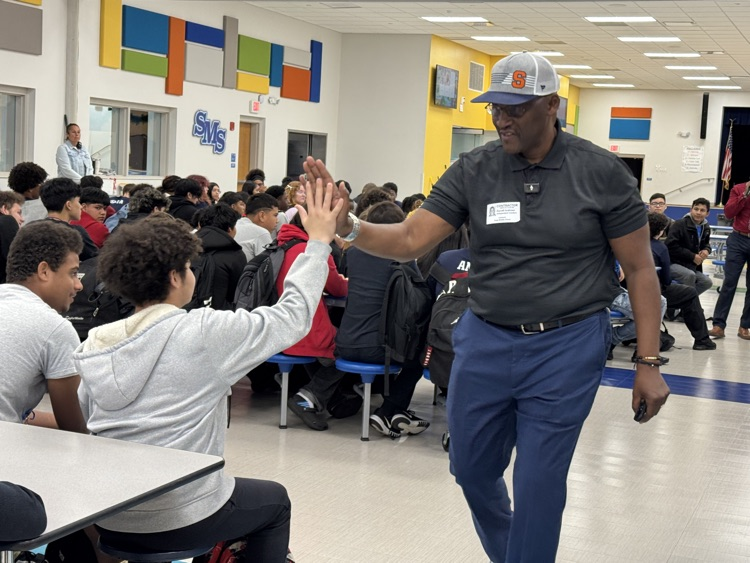a man wearing a ball cap giving a sitting student a high five. 