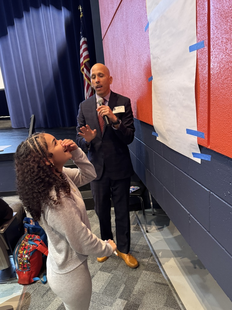 A young woman covers her mouth in astonishment as a man in a suit ask her to jump up higher and draw a line on a sheet of paper taped to the wall