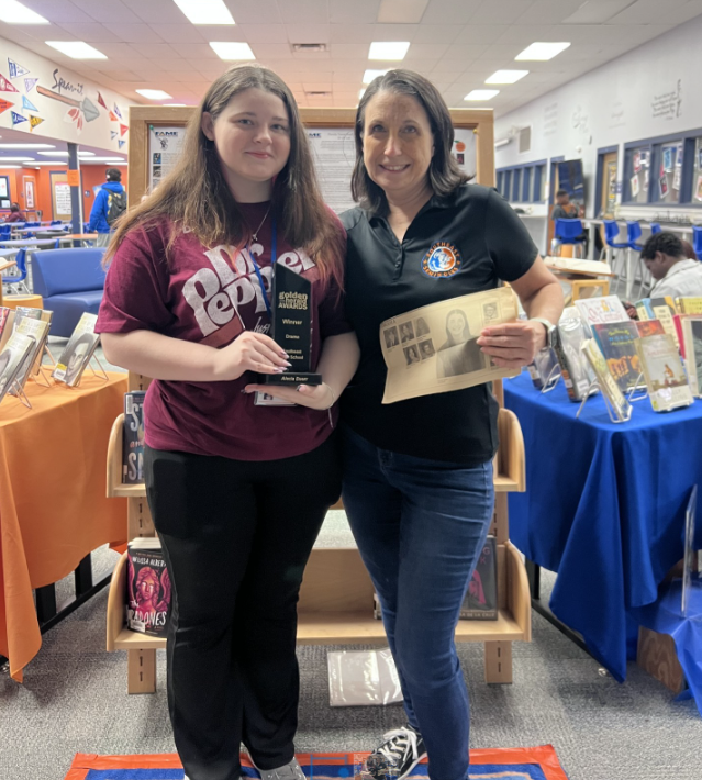 A student wearing a Dr. Pepper t-shirt holding a trophy smiles while standing next to a school media specialist wearing a black polo shirt.