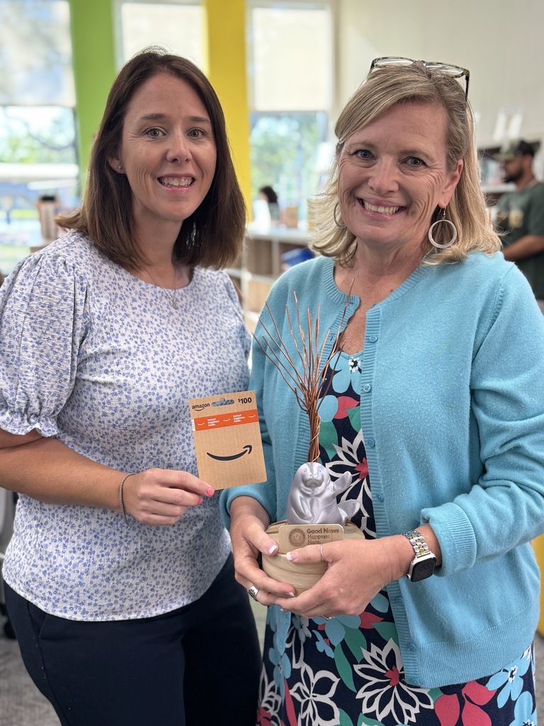 Two women smiling in a school media center. One is holding an Amazon gift card, the other is holding a trophy.