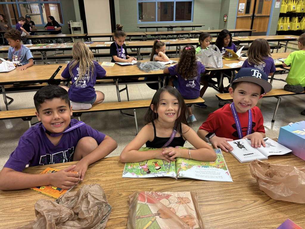 Three children smiling while sitting at a lunchroom table with open books in front of them.