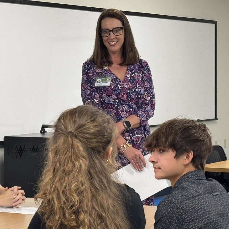superintendent smiles, while standing near a table of seated students speaking