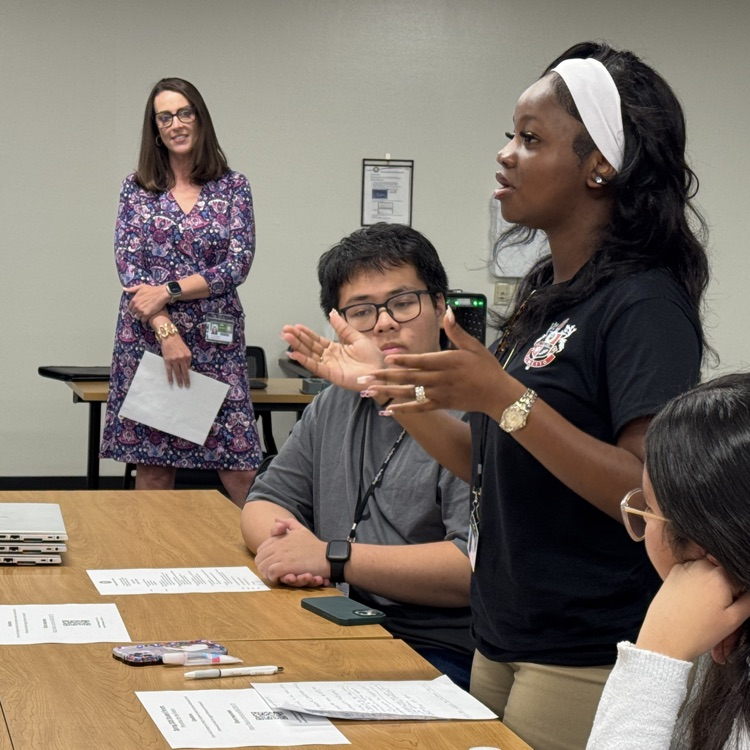 The superintendent of schools looks on as a student standing, speaks to her seated peers