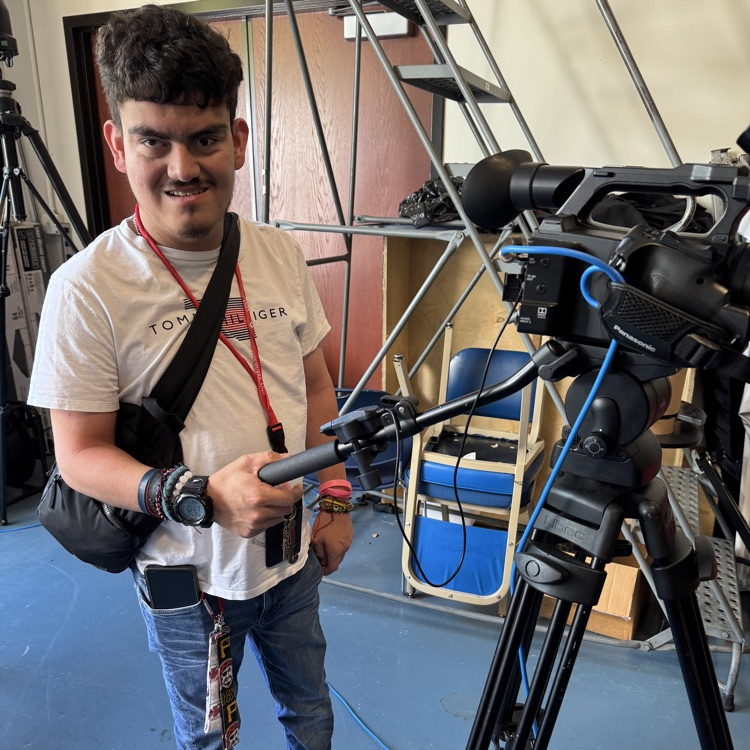A high school student smiles while operating a TV news camera on a tripod
