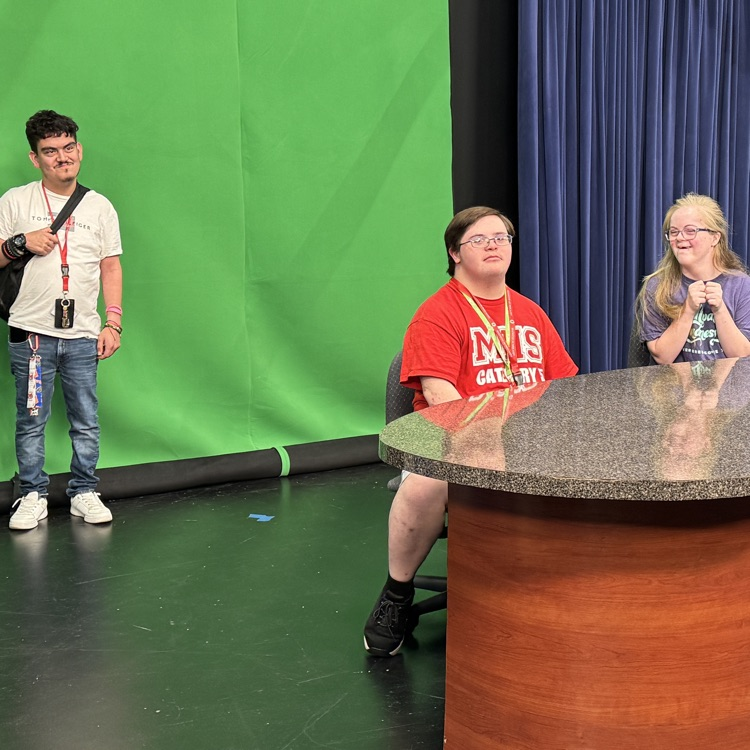 students on a field trip in a television station sitting at a News desk and standing at the green screen