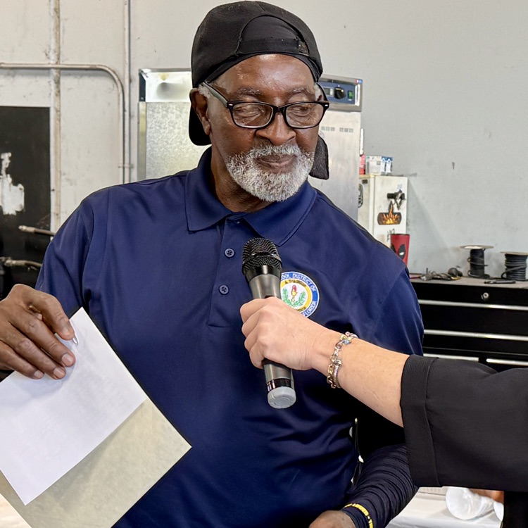 A transportation employee wearing a blue shirt and glasses, about to speak into a microphone while holding a certificate