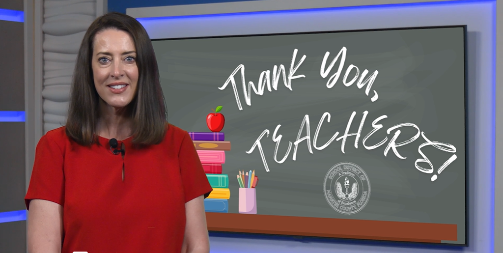 A school district leader wearing a red blouse smiling in front of a chalkboard graphic which reads, "Thank you, Teachers". 
