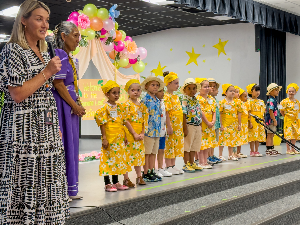 Kindergarten students lined up on stage in yellow costumes as their principal speaks into a microphone.