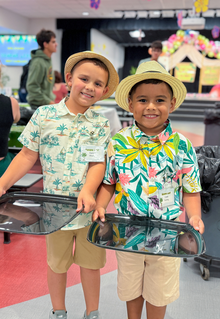 Two boys smiling while wearing tropical shirts and straw hats. They're also holding trays.