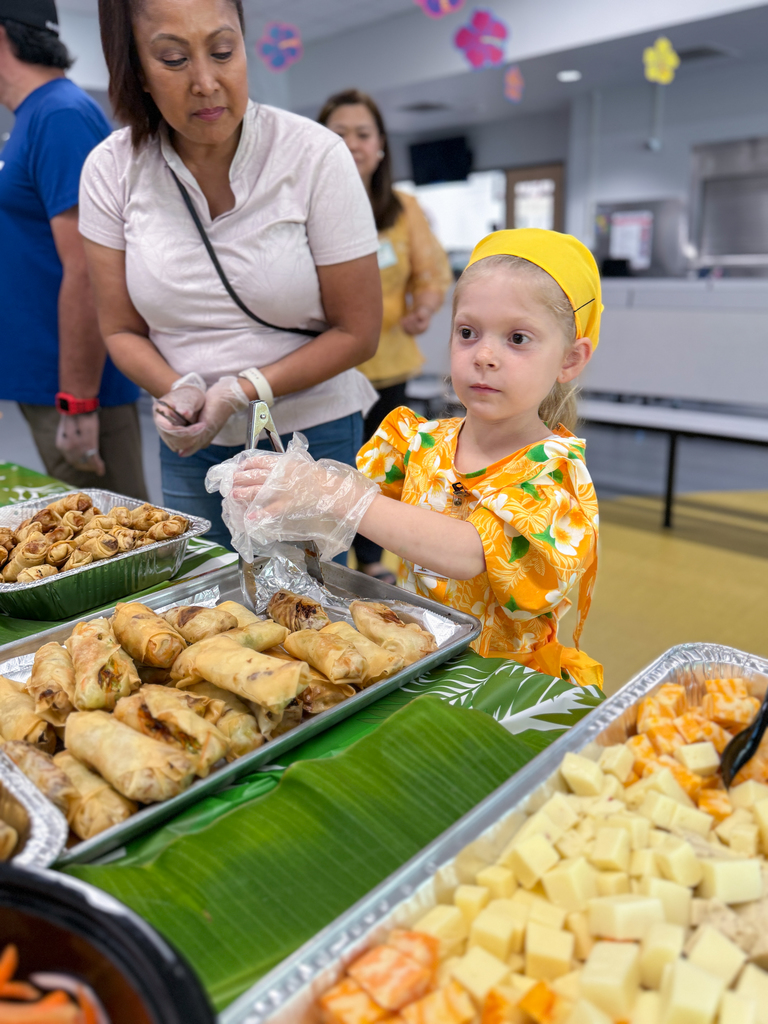 A little girl in a floral yellow dress and wearing gloves helps serve food at a restaurant themed school event.