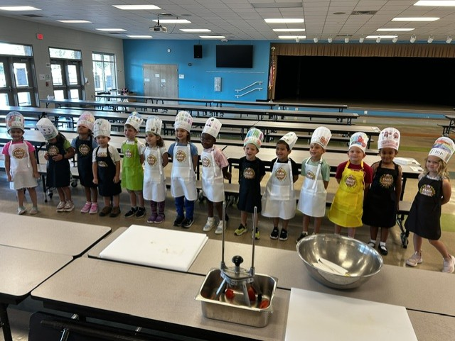 Small children lined up in a cafeteria wearing aprons and chef hats.