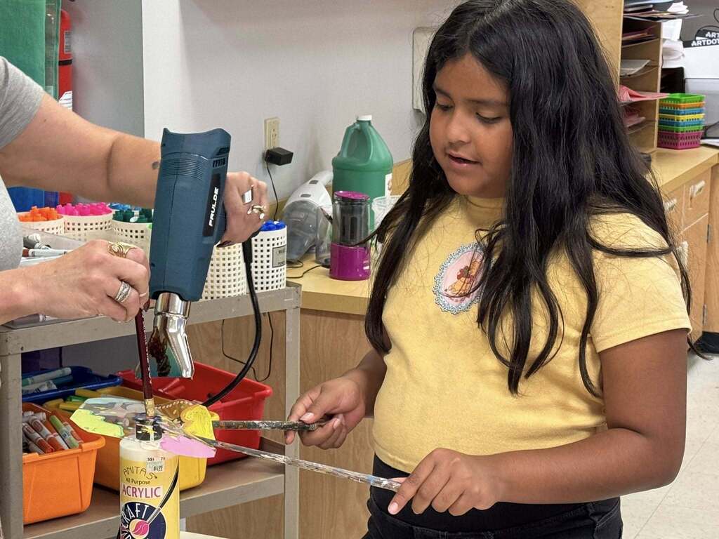 A student wearing a yellow tshirt with long black hair molds a piece of art while holding two wooden rods.