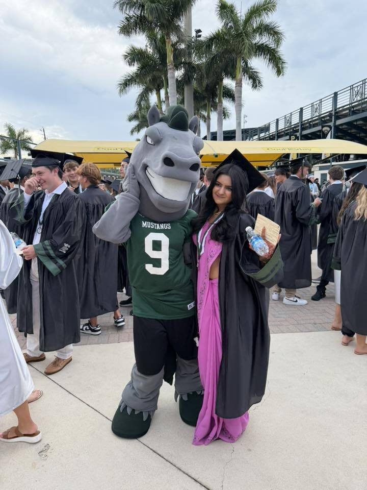 The Mustang horse mascot poses for a photo with a high school graduate wearing a pink dress and black gown.
