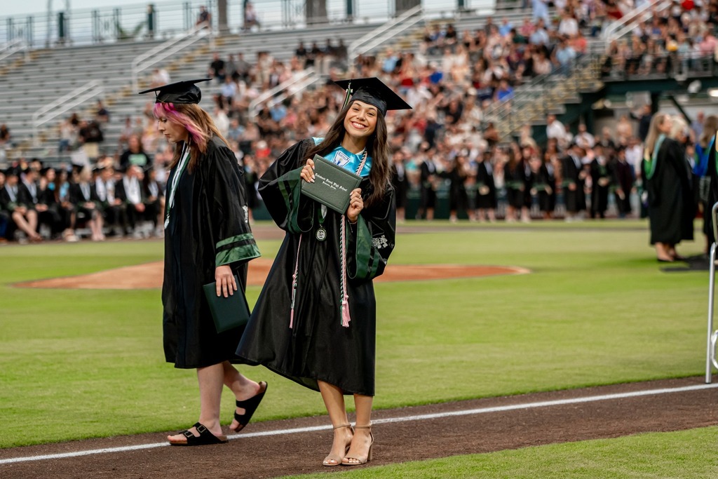 A young woman in heels and black gown holding a high school diploma.
