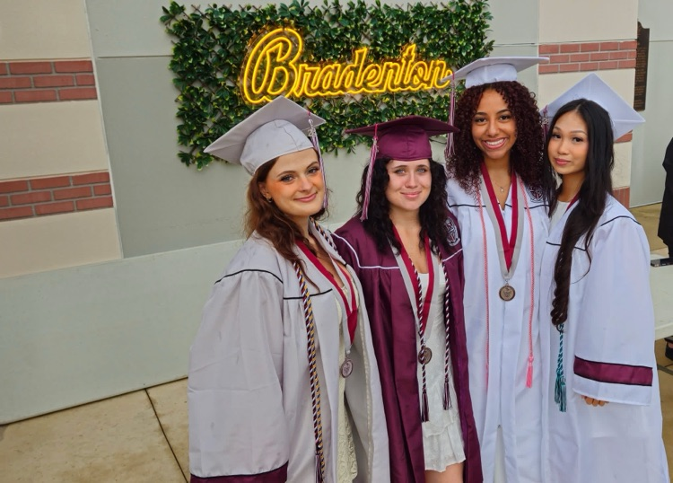 four young women in cap and gown stand in front of a mural that says Bradenton