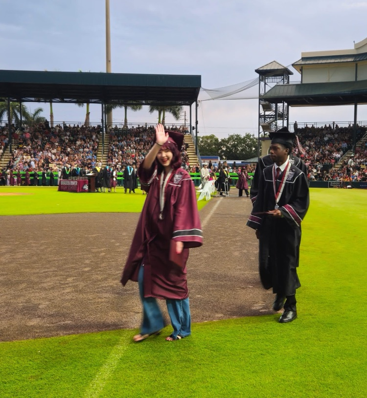 A high school graduate in a maroon cap and gown waves to the audience as she walks across a baseball field