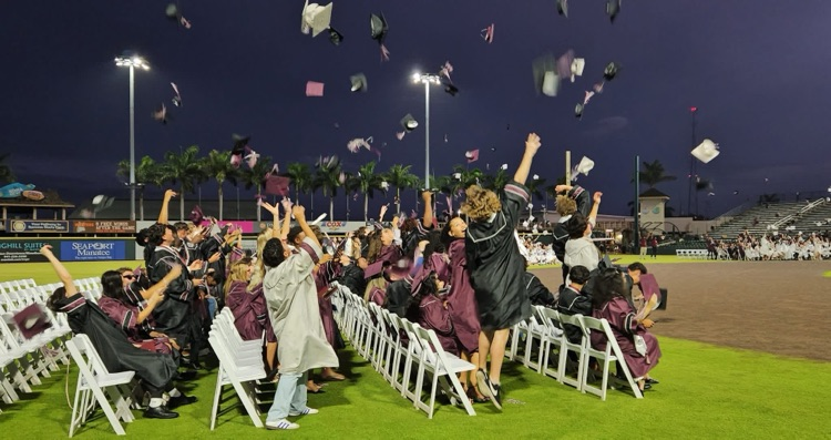 Five rows of high school graduates toss their caps into the air after a ceremony at LECOM Park